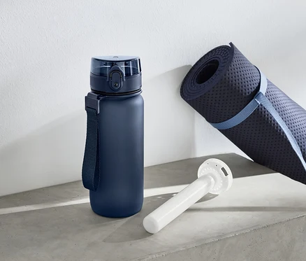 Eine blaue Wasserflasche steht neben einer gerollten Yogamatte und einem weißen, zylinderförmigen Gegenstand.