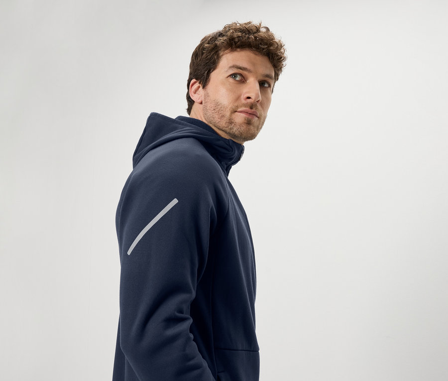 Mann mit braunen lockigen Haaren blickt über die Schulter. Er trägt einen dunkelblauen Kapuzenpullover mit einem diagonalen reflektierenden Streifen am Arm.