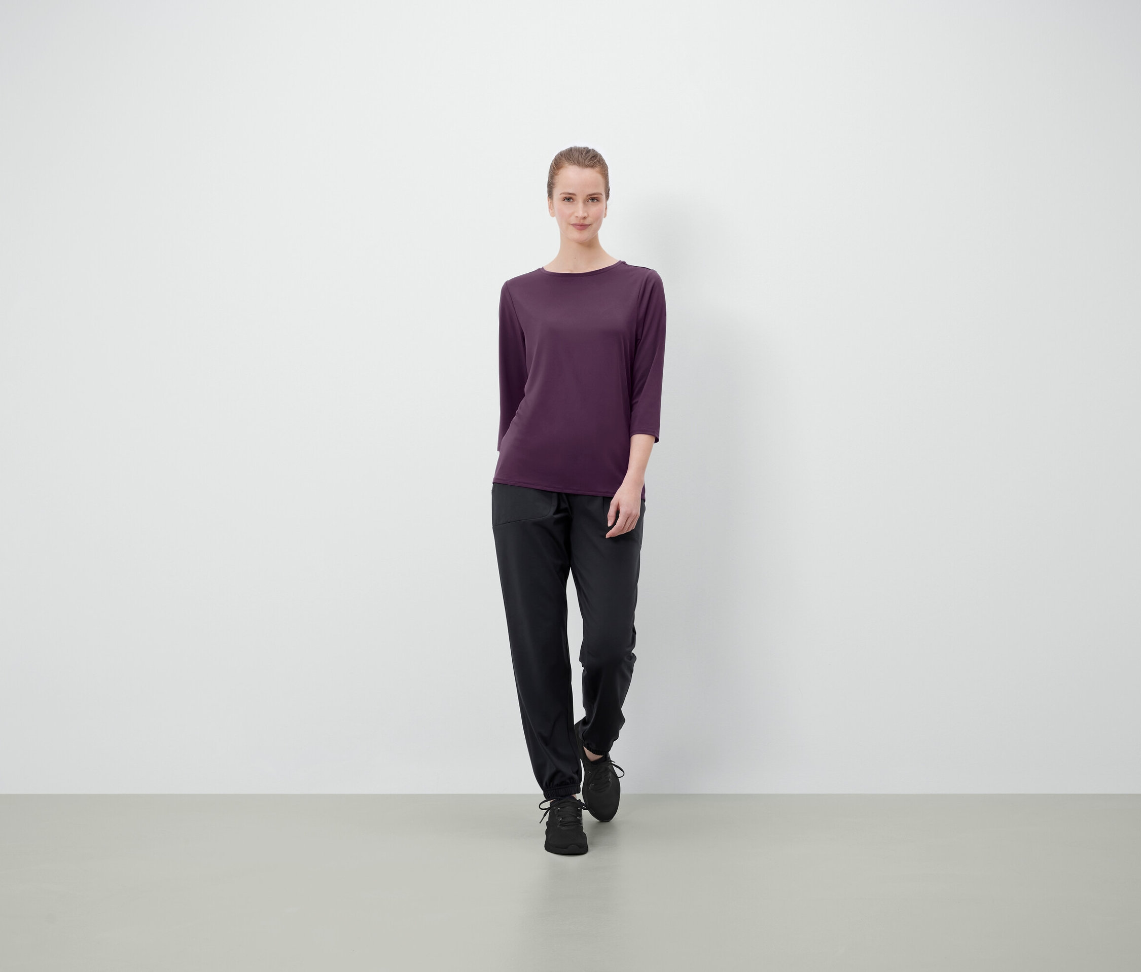 Eine Frau in einem dunkelvioletten Dreiviertelarmshirt, schwarzen Hosen und Schuhen steht vor einem hellen Hintergrund.