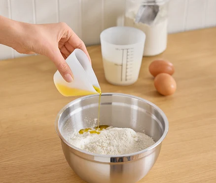 Eine Hand gießt Öl aus einem weißen Messbecher in eine Edelstahlschüssel mit Mehl. Auf der Arbeitsfläche befinden sich zwei Eier, ein Messbecher mit Milch und ein Glas mit Mehl und Löffel.