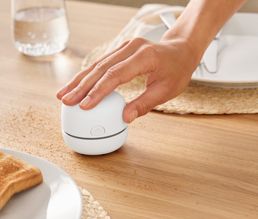 Eine Hand schaltet ein kleines, weißes Gerät auf einem Holztisch neben einem Glas Wasser ein.