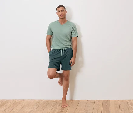 Mann steht auf einem Bein an einer Wand, trägt ein grünes T-Shirt und Shorts.