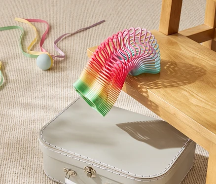 Eine regenbogenfarbene Spielzeugspirale liegt teilweise auf einem beigen Koffer unter einem Holzstuhl.