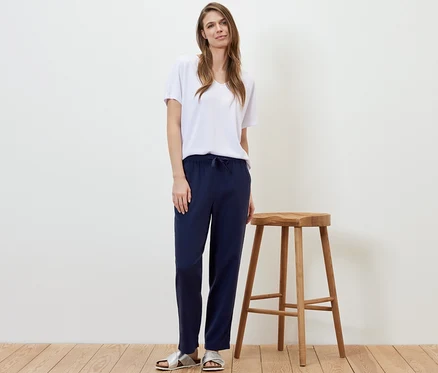 Frau steht neben einem Holzhocker und trägt eine dunkelblaue Leinenhose und ein weißes T-Shirt.