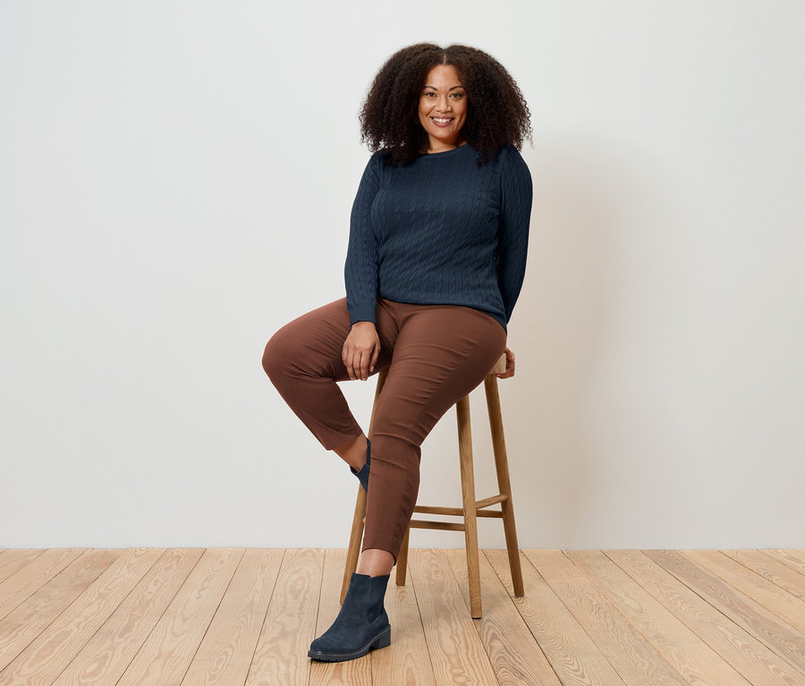 Eine Frau sitzt auf einem Holzhocker und trägt einen dunkelblauen Pullover, braune Hosen und blaue Stiefel.