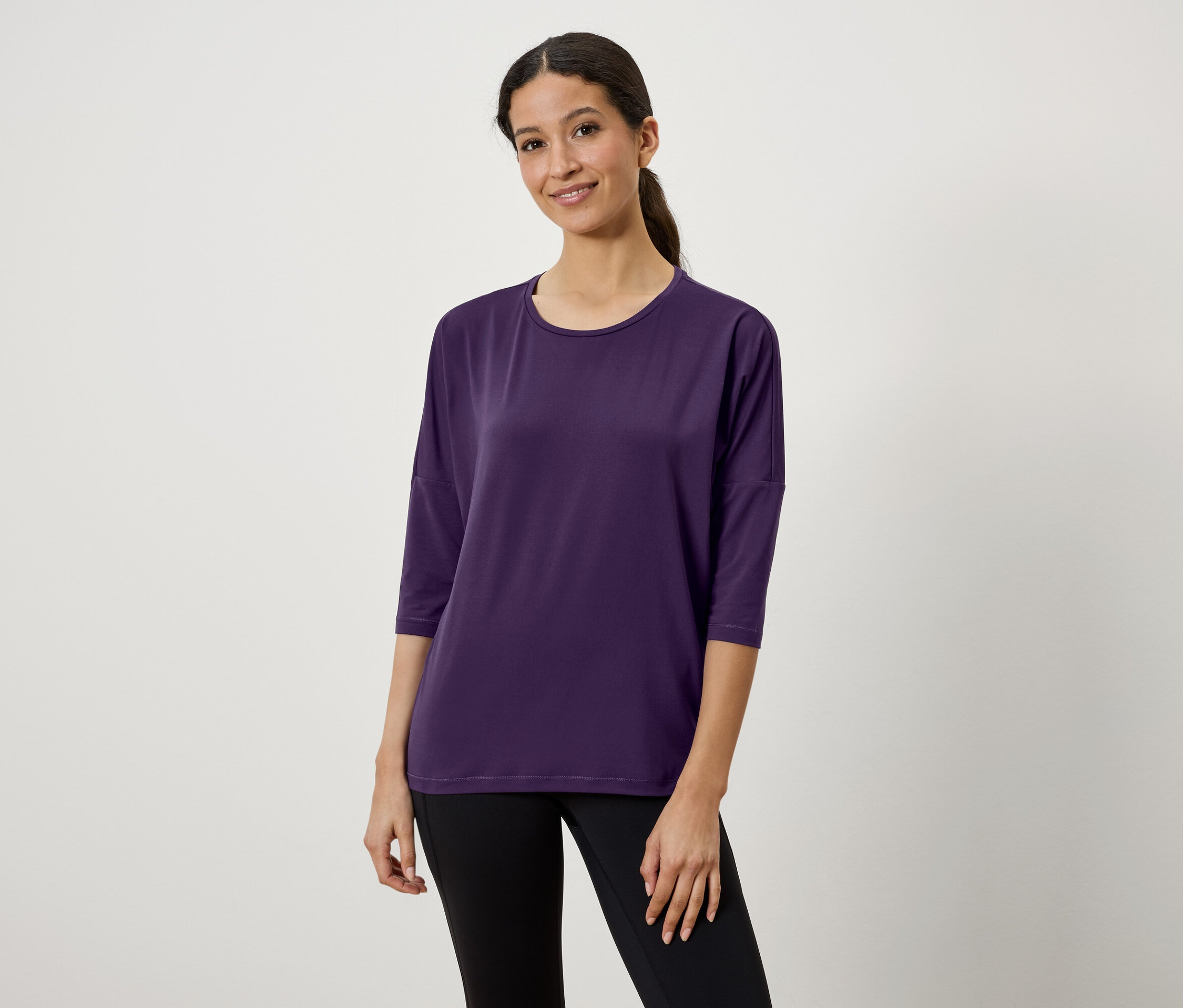 Eine Frau mit braunen Haaren, die zu einem Pferdeschwanz gebunden sind, trägt ein violettes Dreiviertelarm-T-Shirt und schwarze Leggings.