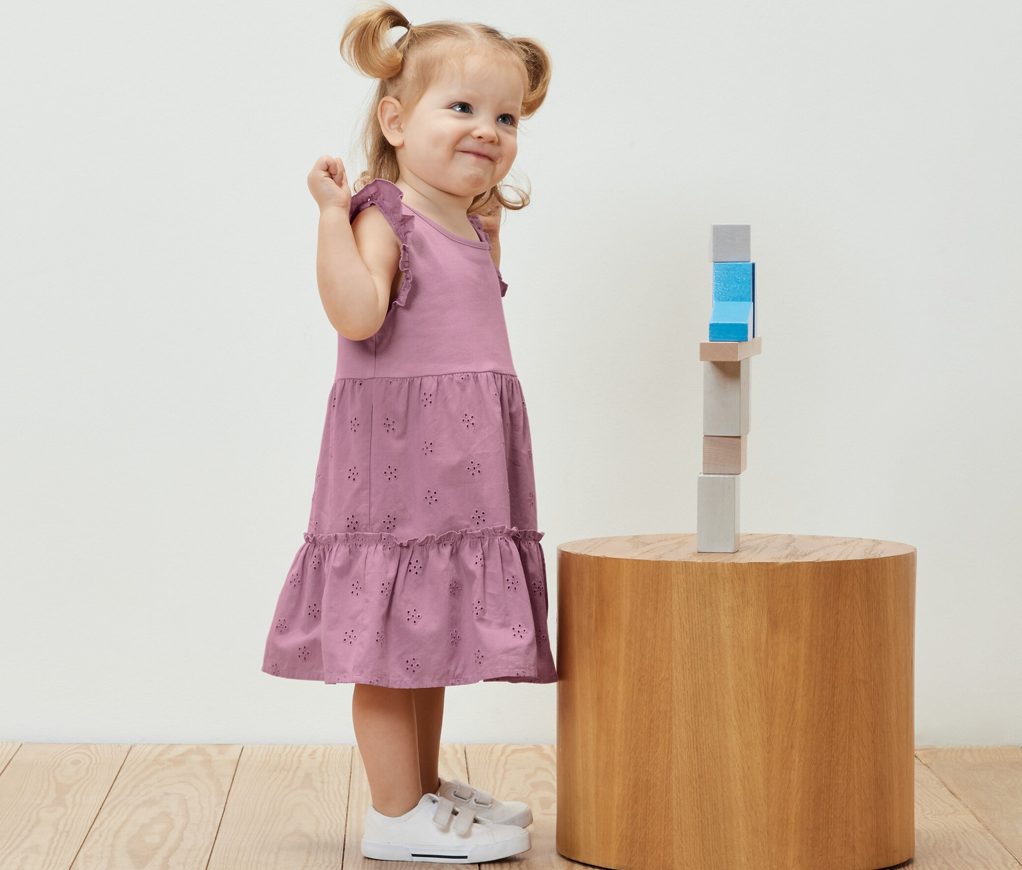 Lächelndes kleines Mädchen im Kinder-Kleid steht neben Holzbausteinen auf einem runden Tisch.
