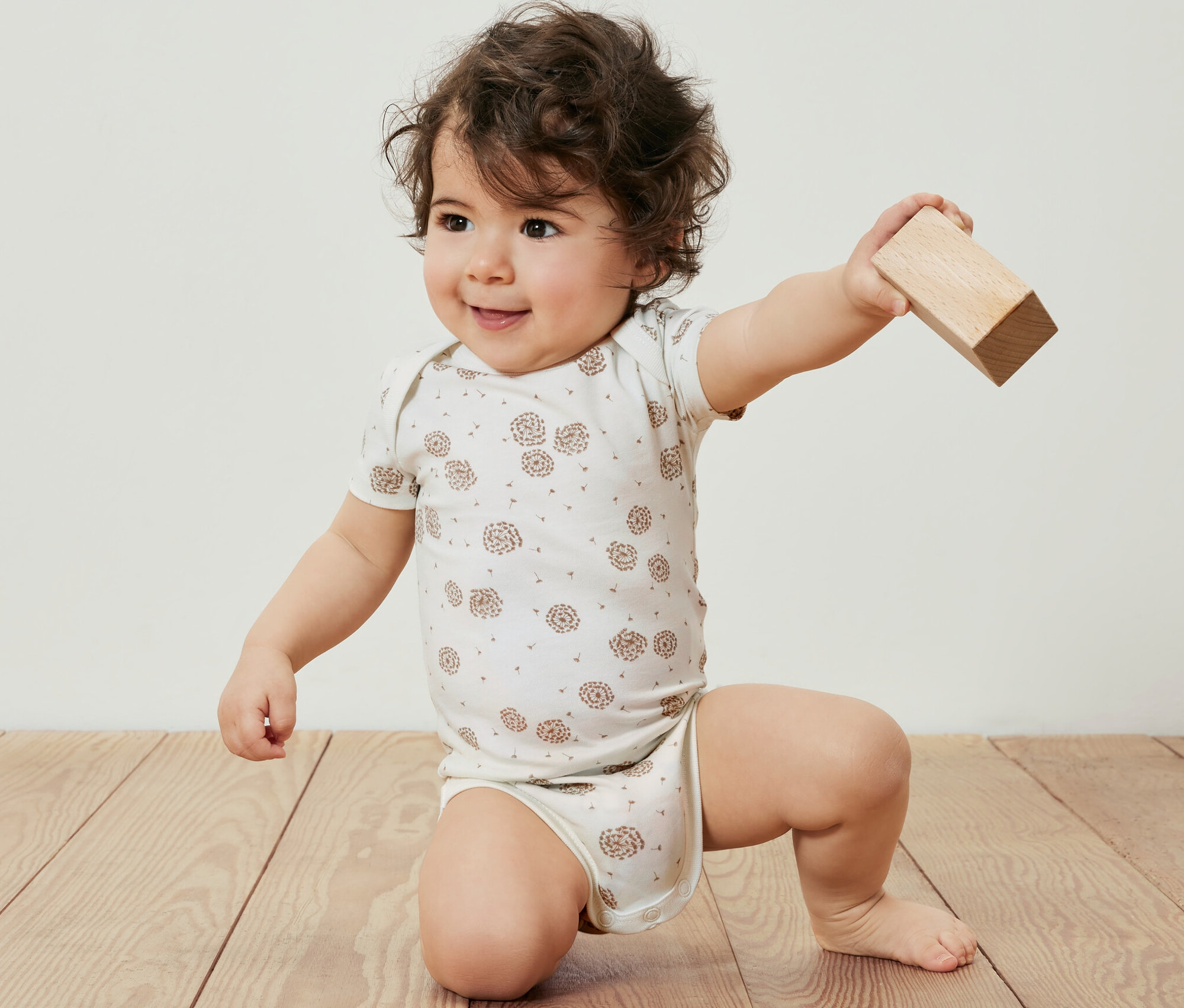 Ein Baby in einem 5er-Pack Baby-Bodys mit Mitwachsfunktion kniet auf einem Holzboden und hält einen Holzblock.