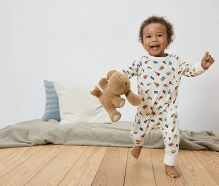Ein lachendes Kleinkind mit lockigem Haar rennt barfuß auf einem Holzboden, einen Teddybären in der Hand und trägt einen weißen Overall mit bunten Automotiven.
