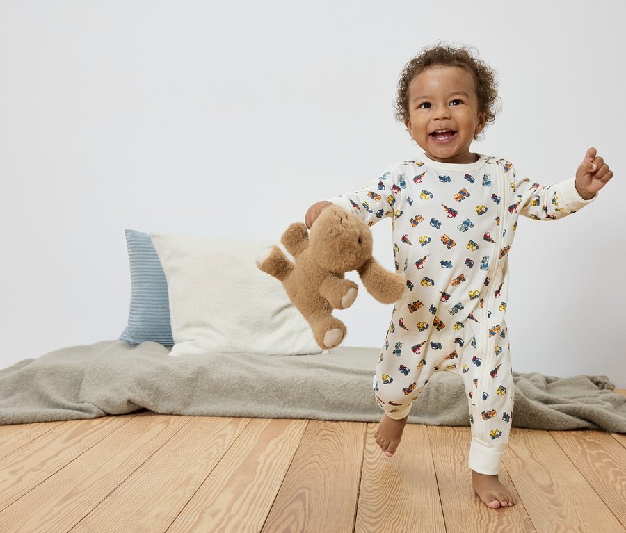 Ein lachendes Kleinkind mit lockigem Haar rennt barfuß auf einem Holzboden, einen Teddybären in der Hand und trägt einen weißen Overall mit bunten Automotiven.
