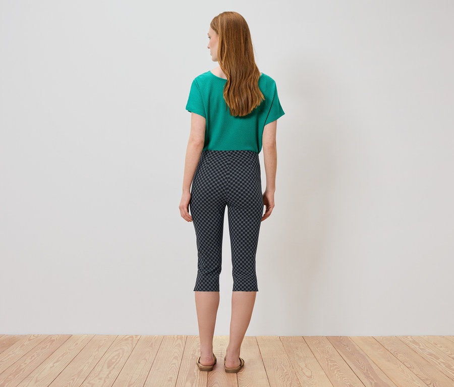 Rückansicht einer Frau mit langen, roten Haaren, die ein grünes T-Shirt, eine gemusterte Dreiviertelhose und Sandalen trägt.