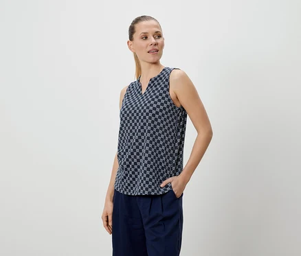 Frau mit blau gemusterter Bluse und blauer Hose, eine Hand in der Hosentasche.