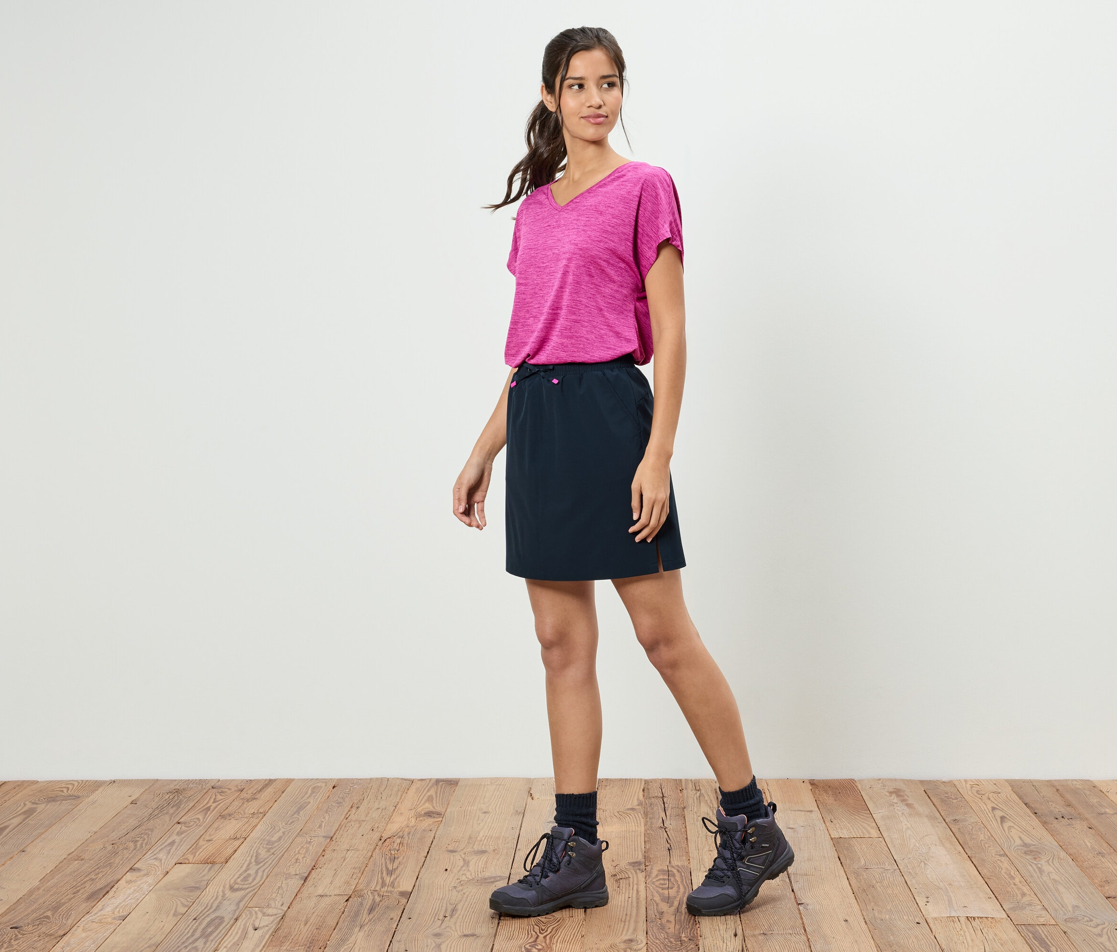 Eine Frau steht vor einer weißen Wand und trägt ein pinkfarbenes T-Shirt, einen schwarzen Rock und Wanderschuhe.
