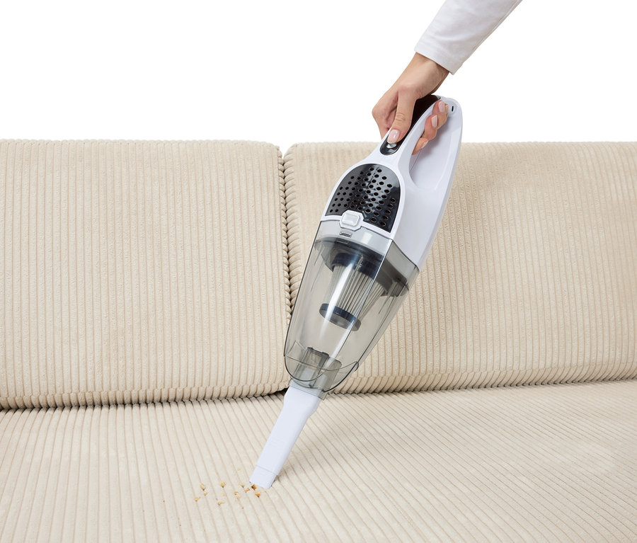 Eine Hand saugt Krümel mit einem 2-in-1-Akku-Staubsauger von einem beigen Sofa.