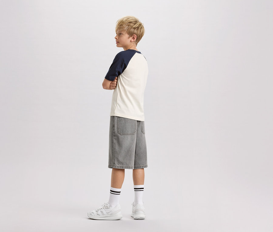Ein blonder Junge steht seitlich, die Arme verschränkt, in einem cremefarbenen T-Shirt mit marineblauen Ärmeln, grauen Jeansshorts, weißen Socken mit schwarzen Streifen und weißen Turnschuhen.