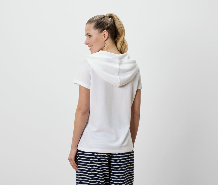 Eine Frau mit blonden Haaren trägt ein weißes Kapuzenshirt und eine blau-weiß gestreifte Hose, von hinten gesehen.