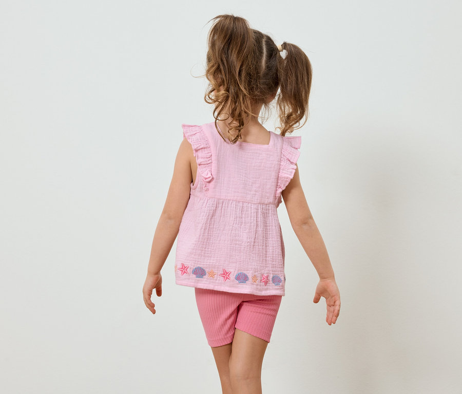 Mädchen von hinten, trägt rosa Top mit Muscheln und Sterne und kurze, enge, rosa Hose vor hellem Hintergrund.