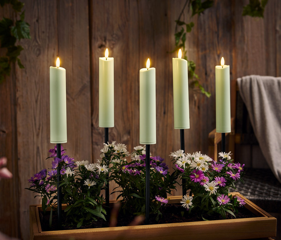 Fünf brennende, grüne Kerzen stehen in einem Holzkasten mit lila und weißen Blumen vor einer Holzwand.