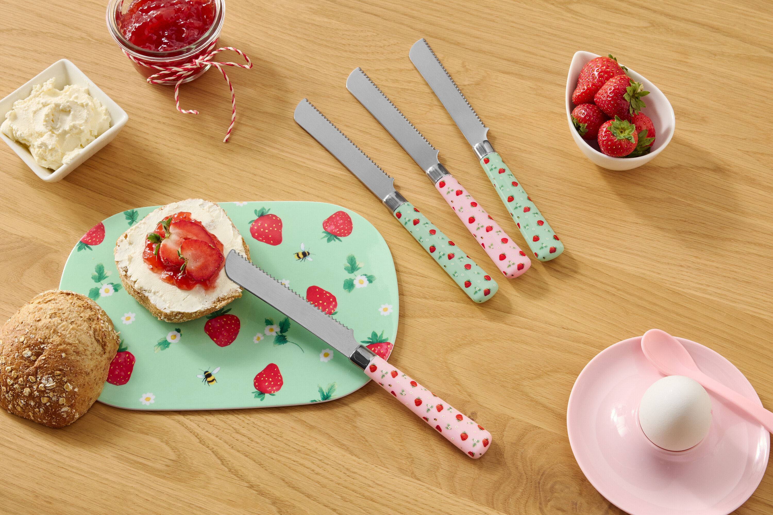 Auf einem Holztisch befinden sich ein Brötchen, ein Teller mit Erdbeermuster, Frischkäse, Marmelade, Erdbeeren und vier Messer mit Erdbeermuster.