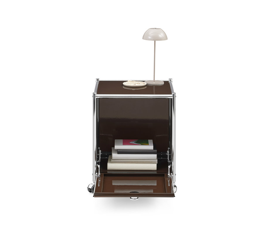 Brauner Modulschrank mit Chromrahmen und Rollen, einer weißen Tischlampe und einer Schale obenauf. Die untere Klapptür ist geöffnet und zeigt Bücher im Inneren.