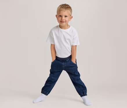 Junger Junge mit blondem Haar, lächelt, trägt ein weißes T-Shirt und dunkelblaue Jogger-Hosen mit den Händen in den Taschen, steht breitbeinig in weißen Socken.