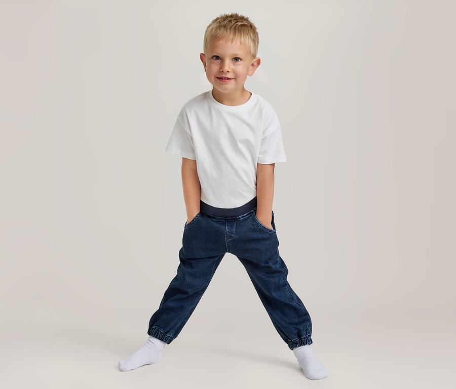 Junger Junge mit blondem Haar, lächelt, trägt ein weißes T-Shirt und dunkelblaue Jogger-Hosen mit den Händen in den Taschen, steht breitbeinig in weißen Socken.