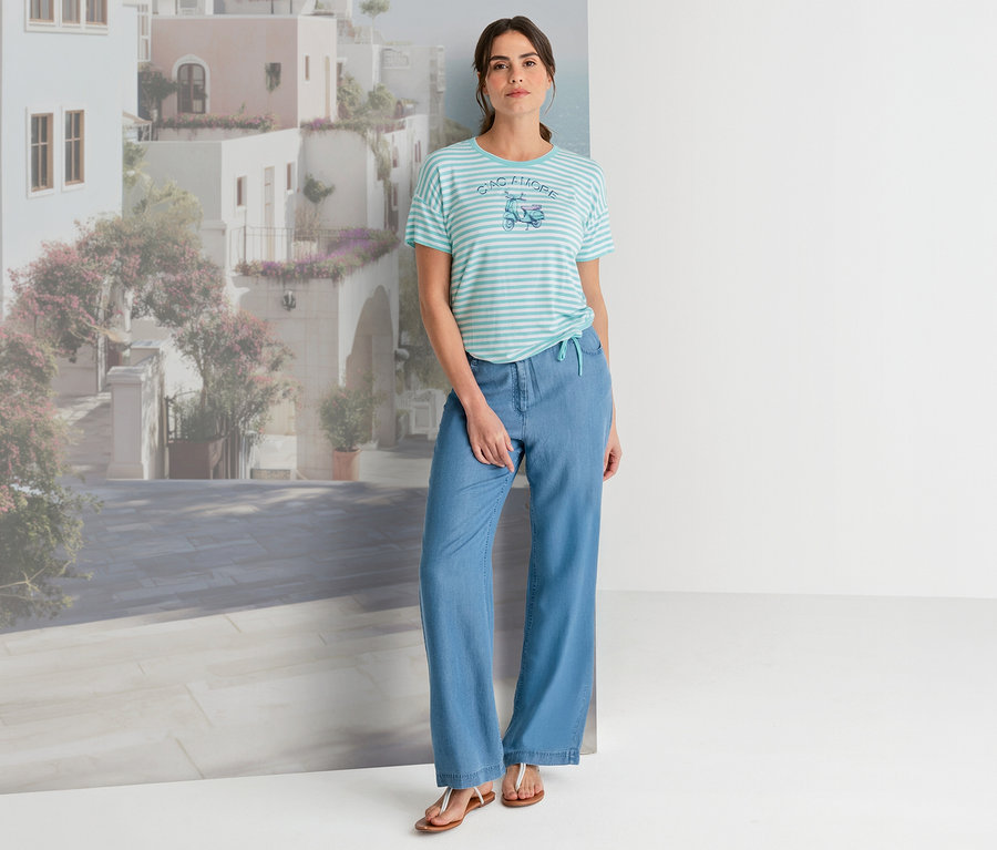 Eine Frau steht vor einer mediterranen Stadtkulisse und trägt ein gestreiftes Olsen T-Shirt mit Roller-Print, hellblaue weite Hosen und braune Sandalen.