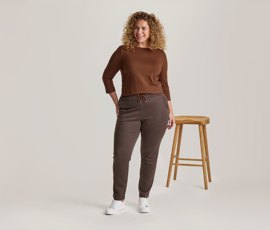 Lächelnde Frau mit lockigem Haar trägt ein braunes Langarmshirt, braune gemusterte Hose und weiße Turnschuhe, steht neben einem Holzhocker.