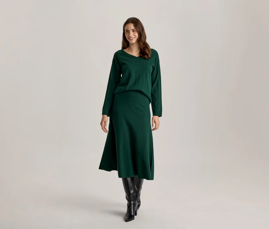 Lächelnde Frau steht, trägt einen dunkelgrünen Strickpullover mit V-Ausschnitt, passenden Midirock und schwarze kniehohe Stiefel.