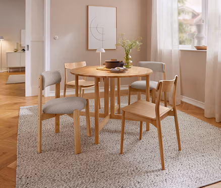 Ein Essbereich mit einem runden Holztisch und vier Holzstühlen auf einem hellen Teppich. Auf dem Tisch stehen eine beige LED-Akku-Tischleuchte, eine Vase mit Pflanzen und ein Topf. Im Hintergrund ist ein Fenster mit Vorhängen und ein Bild zu sehen.