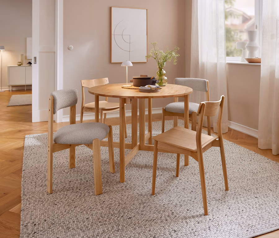 Ein Essbereich mit einem runden Holztisch und vier Holzstühlen auf einem hellen Teppich. Auf dem Tisch stehen eine beige LED-Akku-Tischleuchte, eine Vase mit Pflanzen und ein Topf. Im Hintergrund ist ein Fenster mit Vorhängen und ein Bild zu sehen.