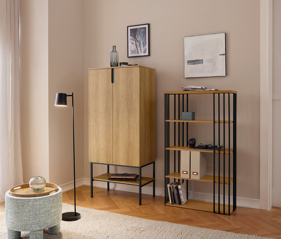 Ein Wohnzimmer mit hellem Holzparkett und beigem Teppich, das einen hellbraunen Schrank, ein Regal mit schwarzen Metallstreben und einen blauen Hocker mit Holztablett zeigt. Eine schwarze Stehlampe steht daneben, und ein Fenster mit Vorhängen ist links sichtbar.