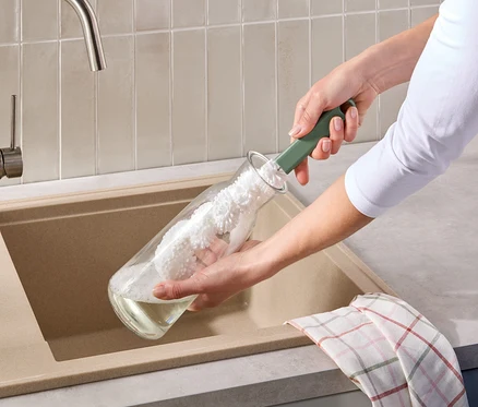 Eine Person reinigt eine Glasflasche mit einer grünen Flaschenbürste über einem beigen Spülbecken. Ein kariertes Geschirrtuch hängt über dem Spülbeckenrand.