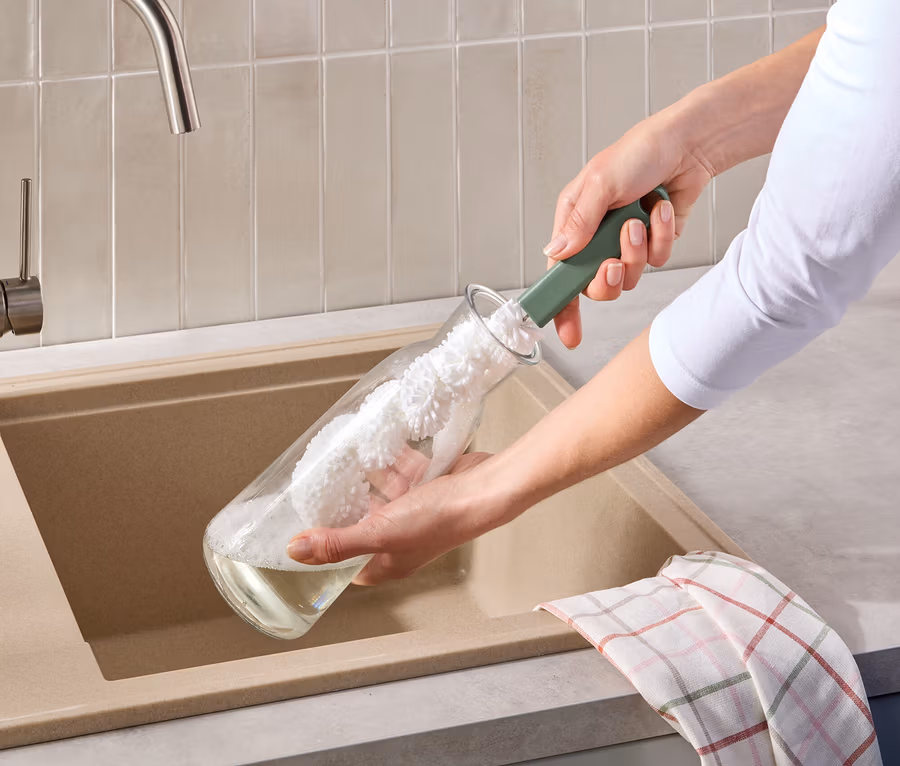 Eine Person reinigt eine Glasflasche mit einer grünen Flaschenbürste über einem beigen Spülbecken. Ein kariertes Geschirrtuch hängt über dem Spülbeckenrand.