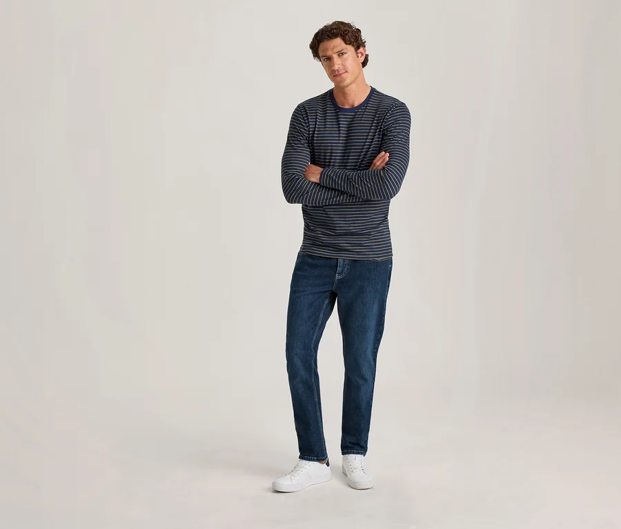 Ein Mann mit lockigem braunem Haar steht mit verschränkten Armen da. Er trägt ein dunkelblaues und hellblaues gestreiftes Langarmshirt, dunkelblaue Jeans und weiße Sneaker vor einem hellen, einfarbigen Hintergrund.