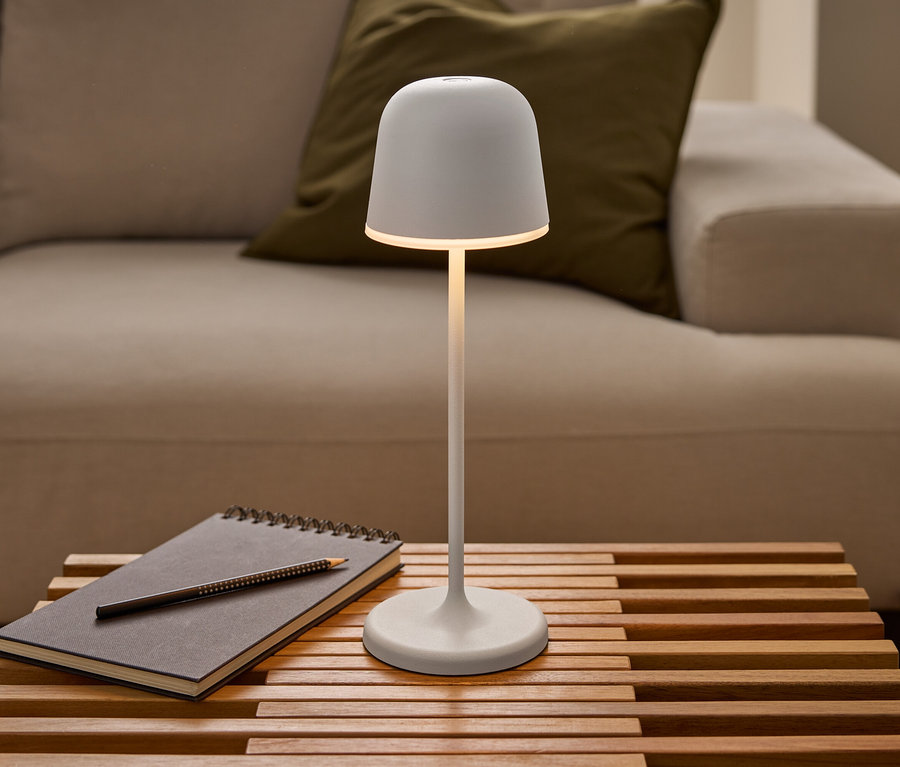 Eine leuchtende weiße Eglo LED-Akku-Tischleuchte »Mannera« steht auf einem Lattenholztisch neben einem Notizbuch mit Bleistift vor einem Sofa.