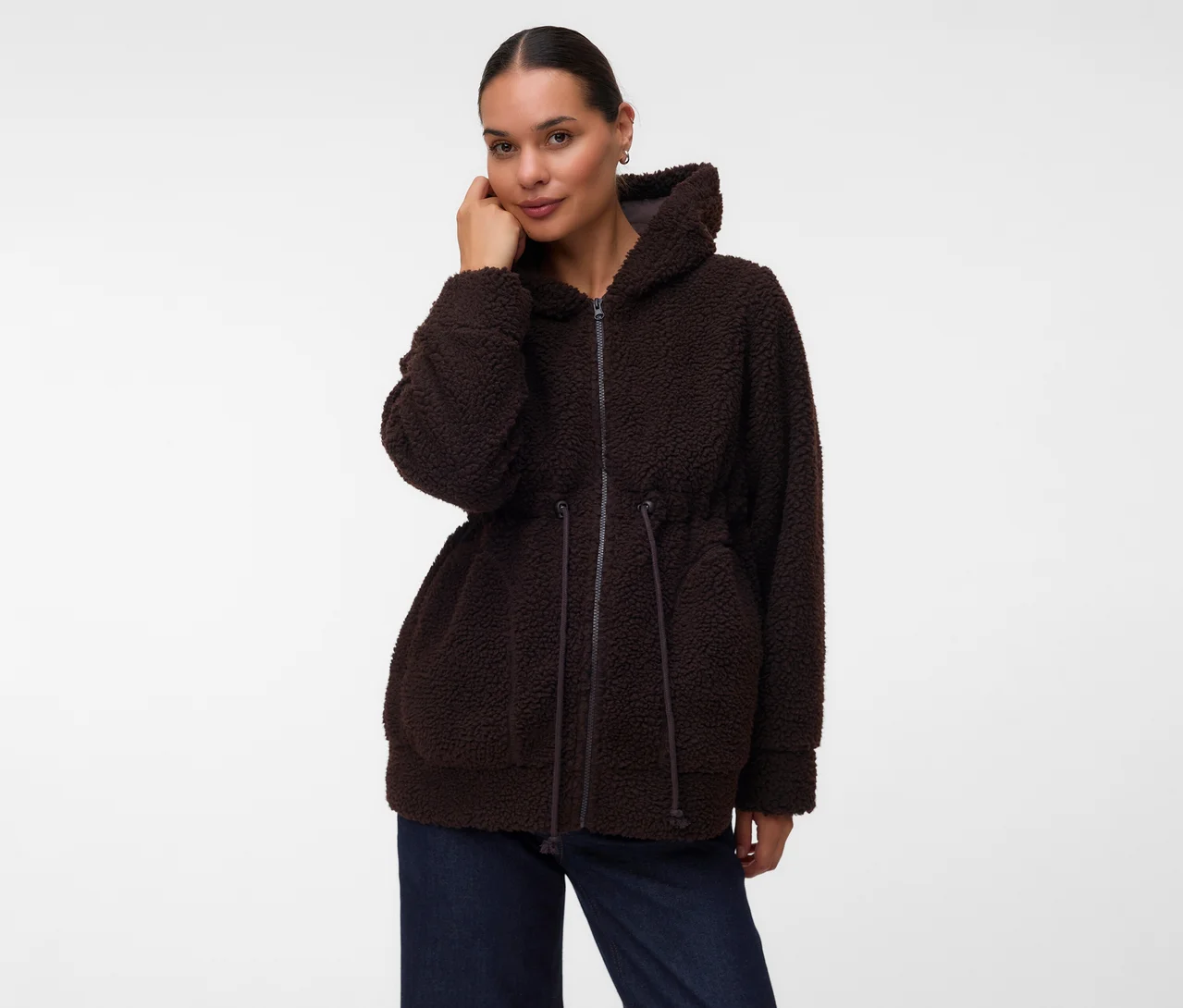 Frau trägt eine dunkelbraune, flauschige Kapuzenjacke mit Reißverschluss und Kordelzug in der Taille, kombiniert mit dunkelblauen Jeans. Sie blickt in die Kamera, eine Hand am Gesicht.