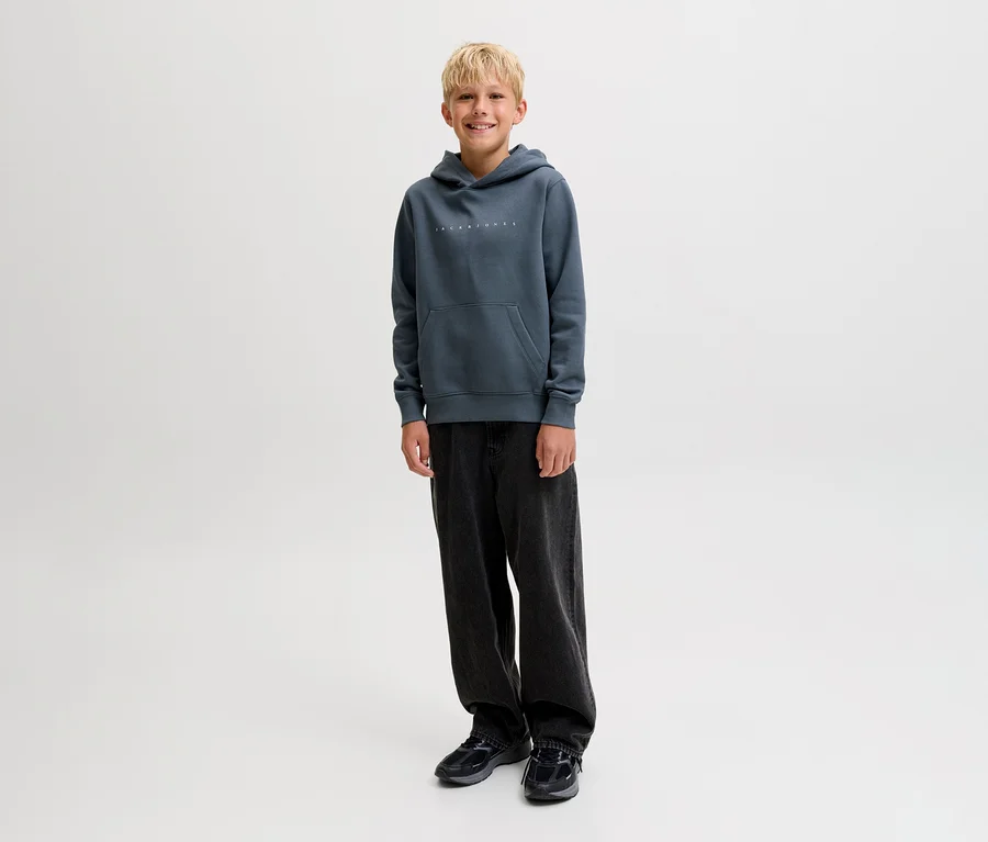 Ein lächelnder Junge steht in einem dunkelgrauen Jack&Jones Star Sweat-Hoodie und einer weiten, dunkelgrauen Hose.