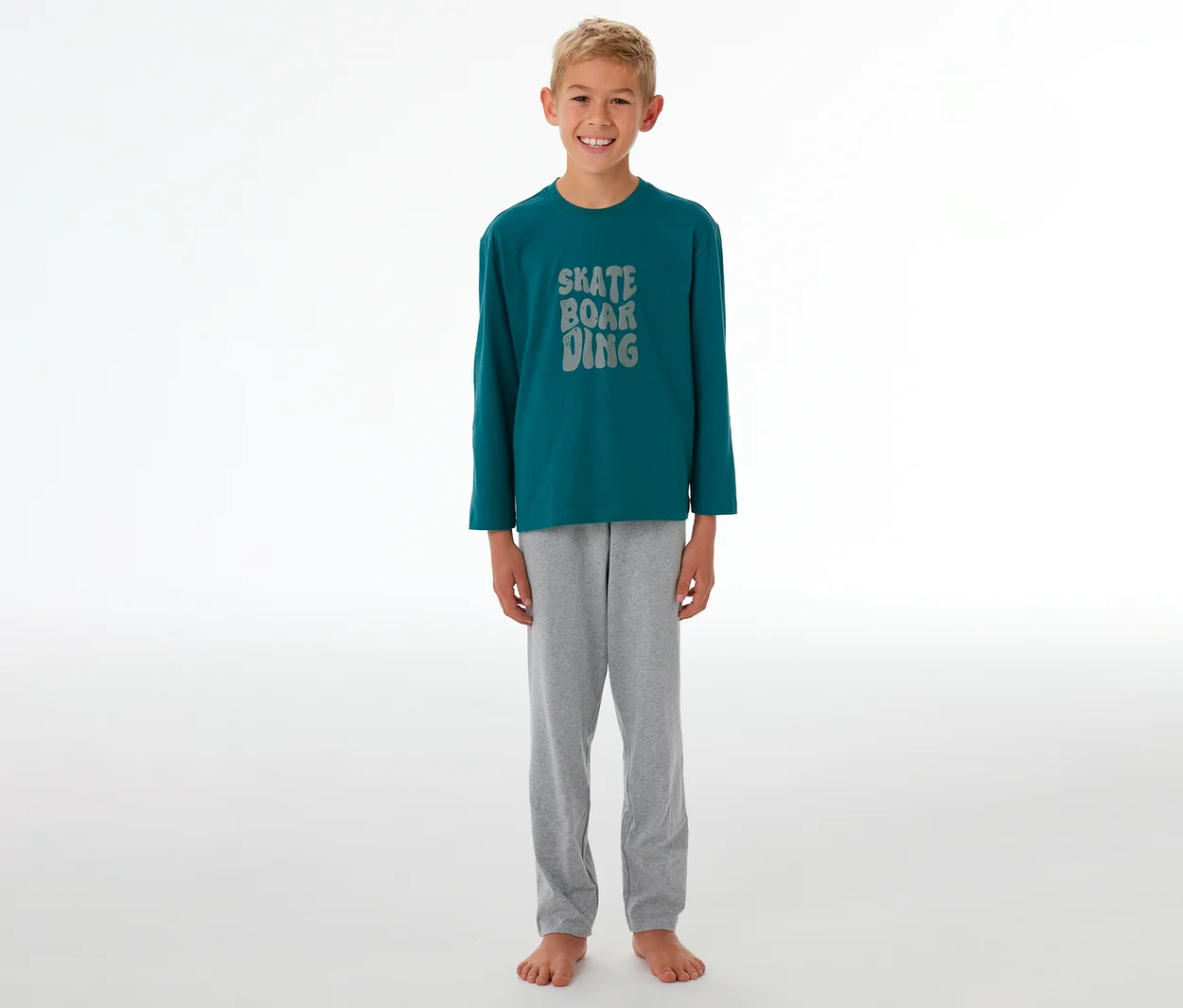 Lächelnder Junge in einem Kinder-Pyjama mit petrolfarbenem Langarmshirt, grauer "SKATE BOARDING"-Aufschrift und grauer Pyjamahose.