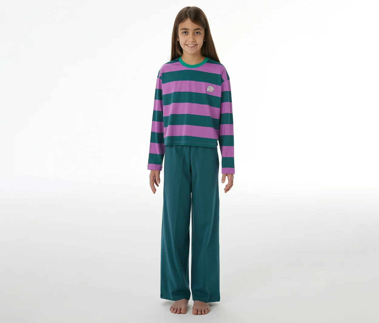 Mädchen lächelt, trägt einen SCHIESSER Kinder-Pyjama mit lila-grün gestreiftem Langarmshirt, grüner Hose und hat nackte Füße.