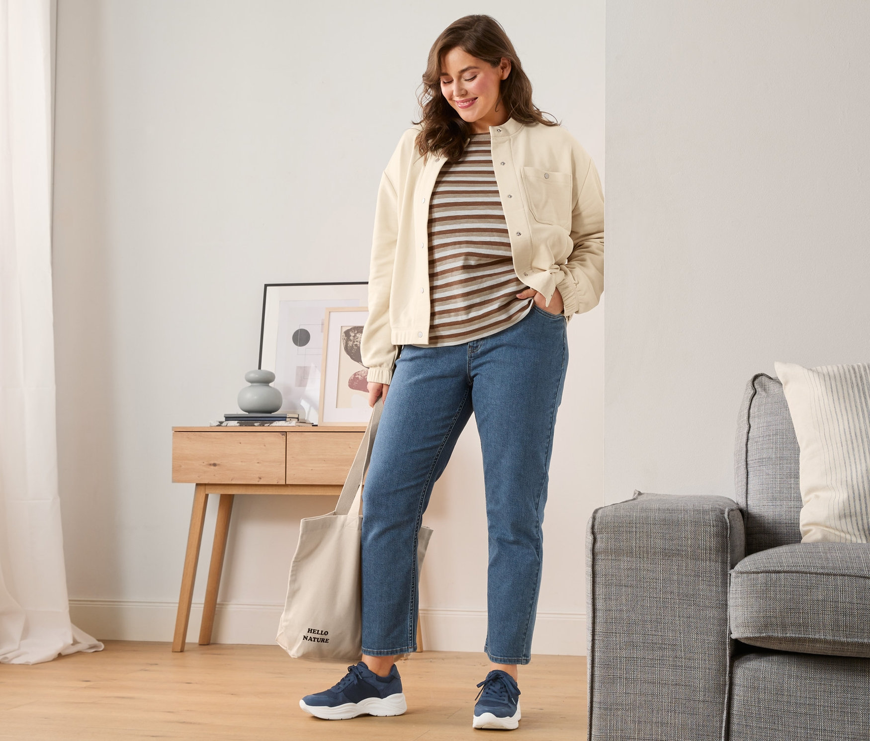 Eine Frau steht im Wohnzimmer und trägt Leder-Sneaker, Straightfit-Jeans, ein braunes, gestreiftes Langarmshirt und eine Sweatjacke. Sie hat einen Canvas-Shopper über der Schulter.