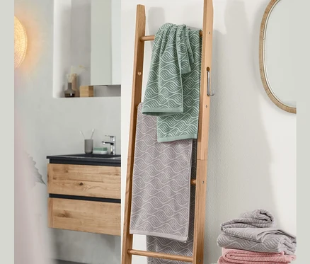Im Badezimmer steht eine Kleiderleiter aus Eiche, an der ein Jacquard-Badetuch und 2 Jacquard-Handtücher hängen. Neben der Leiter befindet sich ein Stapel Jacquard-Handtücher und Badetücher sowie ein Doppelwaschtisch inkl. Mineralgusswaschbecken.