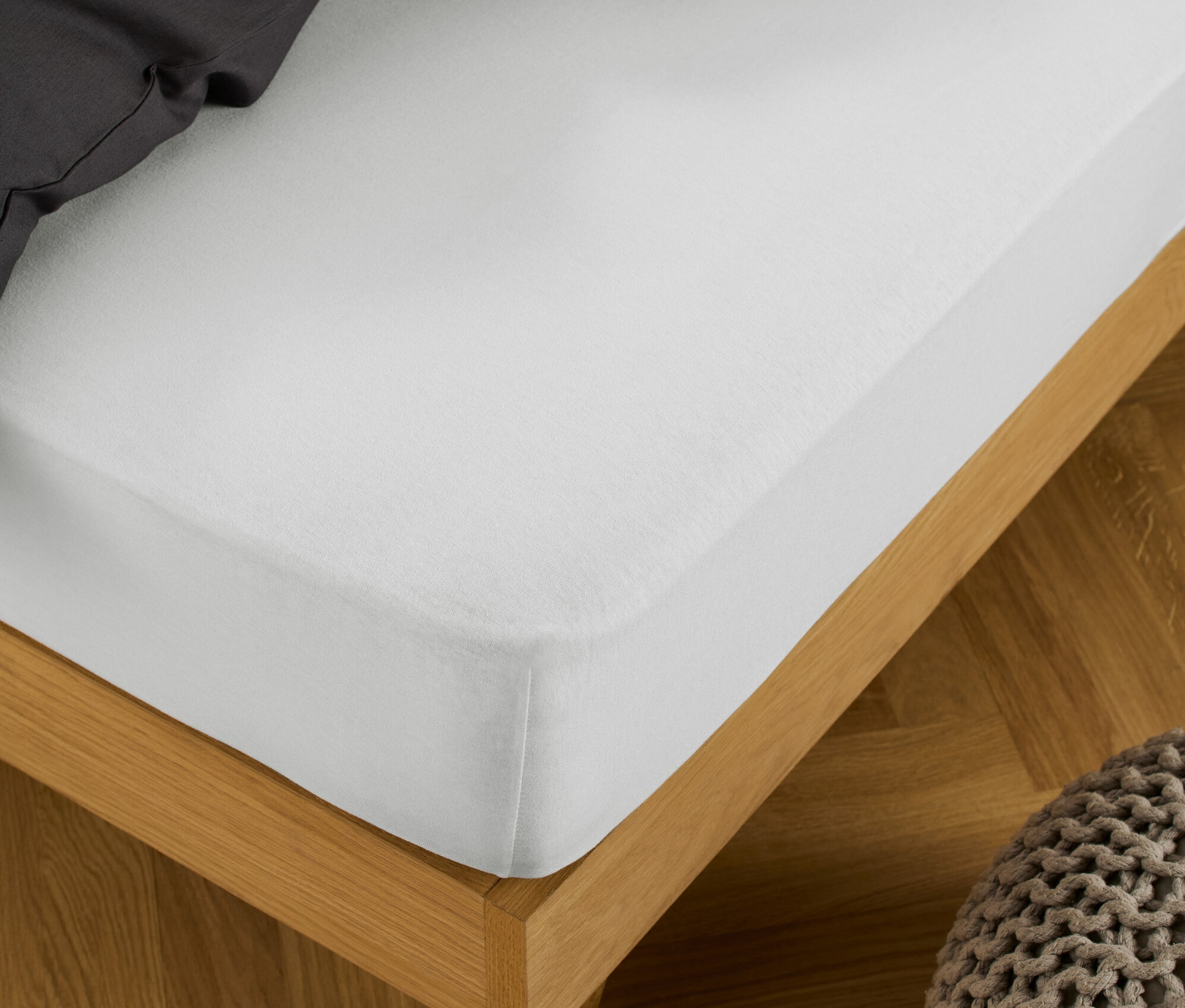 Ein weißes Jersey-Spannbettlaken ist auf ein Bett mit Holzrahmen gespannt, daneben steht ein Strickhocker.
