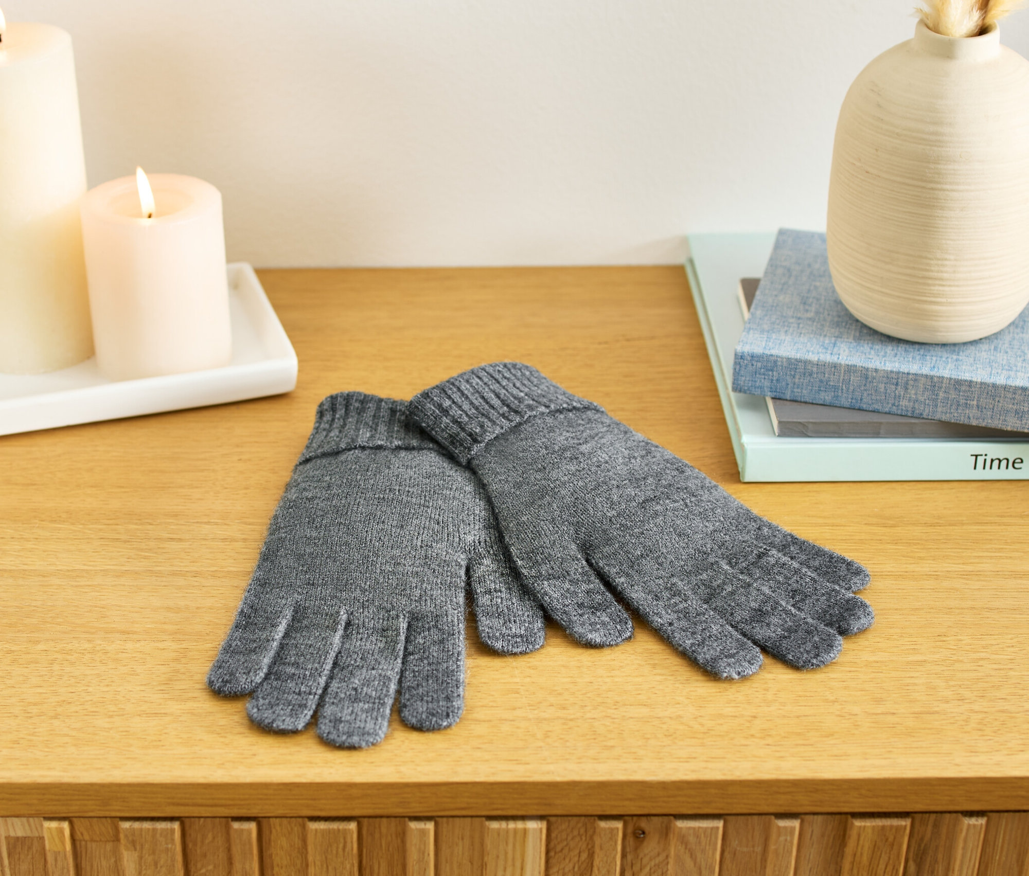 Auf einer Holzkommode liegen grau melierte Strickhandschuhe. Im Hintergrund sind zwei Kerzen, eine Vase und Bücher zu sehen.