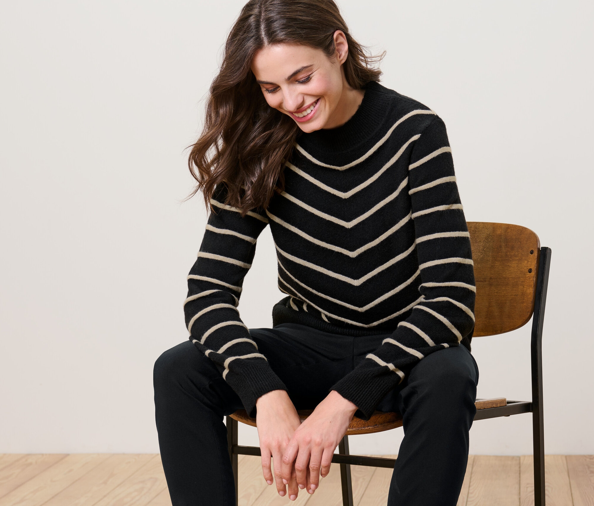Frau sitzt auf einem Stuhl und trägt einen gestreiften Strickpullover mit Stehkragen, schwarz.