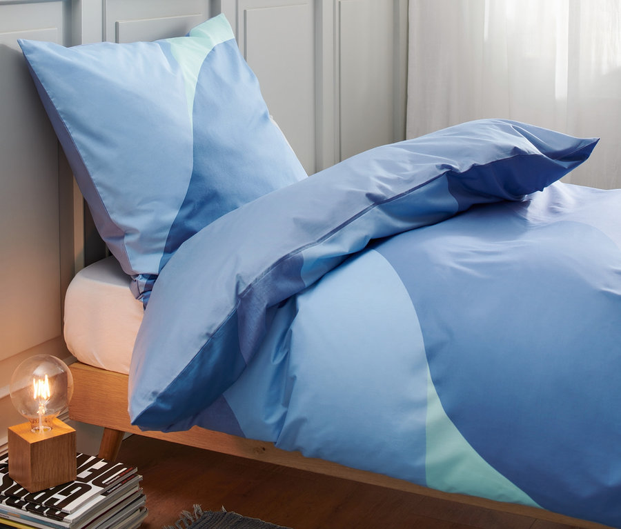 Blaue Premium-Baumwoll-Bettwäsche in Standard- und Übergröße auf einem Bett. Im Hintergrund eine Lampe und Dekoration.