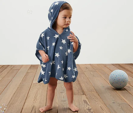 Ein Kind steht auf einem Holzboden und trägt einen blauen Frottierponcho mit Kapuze und weißen Sternen.