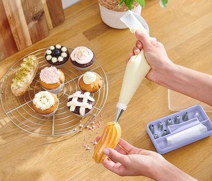 Eine Person dekoriert ein Eclair mit Creme, wobei sie ein Spritztüllen-Set verwendet. Auf dem Tisch liegen Muffins und eine Schale mit Früchten.