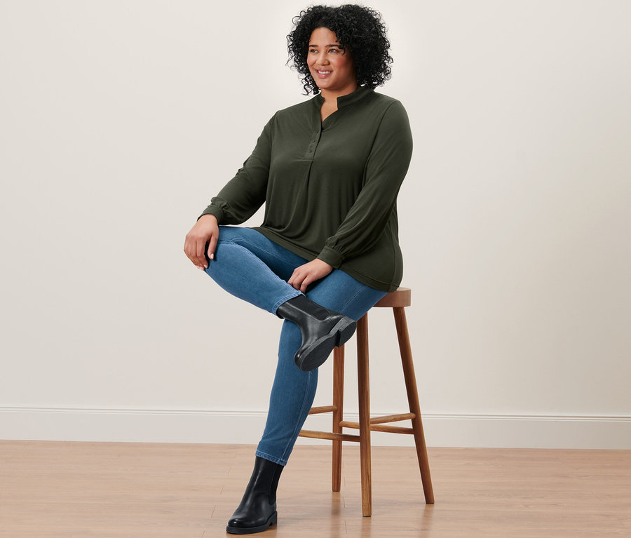Frau sitzt auf einem Hocker und trägt Jeggings, Lederboots und ein Blusenshirt.