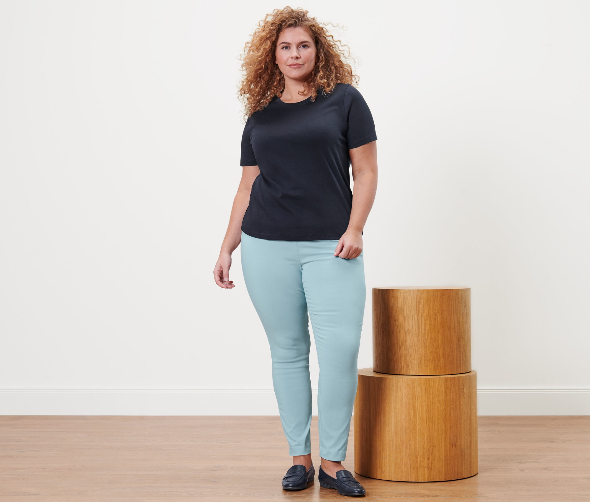 Frau steht und trägt ein dunkelblaues Basic T-Shirt, eine Stretchhose und Leder-Loafer.